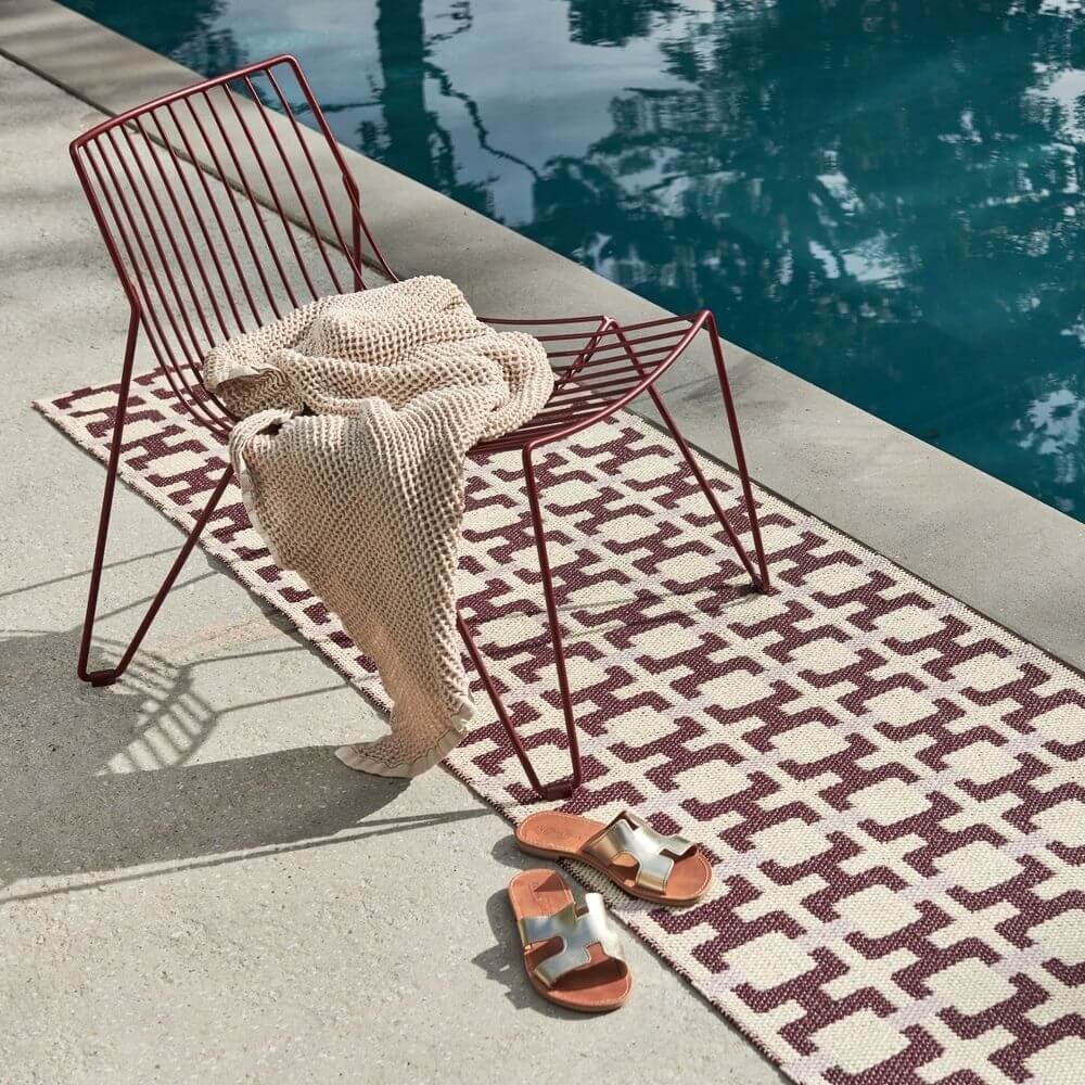 Red metal chair with a beige towel and sandals on a Pappelina rug by a pool.